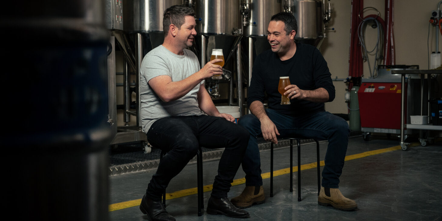 Brenton Fischer and Andrew Forster sitting on stools holding beers
